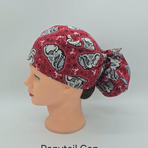 May include: A red ponytail cap featuring a repeating pattern of grey and white elephant illustrations and small white script logos. The fabric has a dark red background with black accents and a gathered back section for hair containment.