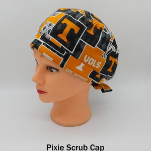 May include: A patterned pixie scrub cap with orange and black "T" letters and the word "VOLS". The cap is displayed on a mannequin head. The text "Pixie Scrub Cap" is at the bottom of the image.