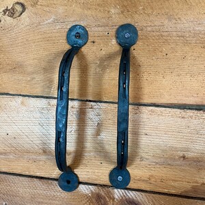 May include: Two dark gray, hand-forged metal door handles featuring a textured, hammered finish. Each handle is curved and secured at both ends by circular mounting plates with visible screws, creating a sturdy and functional hardware set.