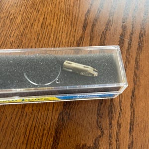 May include: A small, metallic, rectangular cartridge component housed inside a clear plastic storage case with black foam padding. The case features a yellow and blue label with the partial text "Electro-Voice" and "CARTRIDGE" printed on the side.