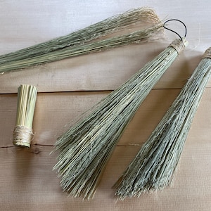 May include: A collection of handmade brooms made from natural materials. The brooms have long, green bristles and are bound with twine. One broom has a black loop for hanging. The brooms are arranged on a light brown wooden surface.