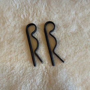 May include: Two black cotter pins, or split pins, are shown against a cream-coloured, textured background. These metal fasteners have a distinctive looped head and two prongs.