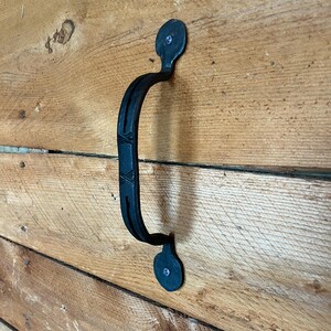 May include: A black metal door handle with a curved, textured grip and two circular mounting plates, each secured with a single visible screw. The handle features a central decorative cross-hatch pattern and is mounted vertically on a wooden surface.