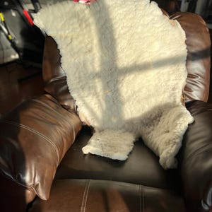 以下が含まれることがあります： 白い羊の毛皮の敷物が、茶色の革製のゆったりとした椅子の上に置かれています。敷物は柔らかくふわふわしていて、椅子は快適でくつろぎやすいです。