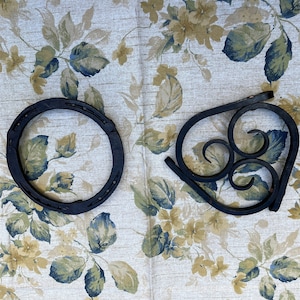 May include: Two black metal decorative items on a floral fabric background. One is a horseshoe-shaped ring and the other is a heart-shaped design with swirls.