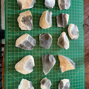 May include: A collection of 14 grey, brown, and white flint arrowheads on a green cutting mat with a ruler.