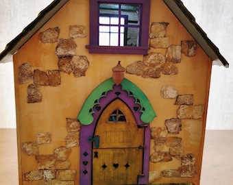 May include: A whimsical wooden fairy house with a stone-like exterior, a purple-framed window, and a green and purple arched doorway. The house features a wooden door, an acorn detail, and sits on a wooden base with a painted green lawn.