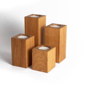 Könnte beinhalten: Vier hölzerne Kerzenhalter unterschiedlicher Höhe. Jeder Halter ist ein quadratischer Block mit einer vertieften Oberseite, um eine weiße Kerze aufzunehmen. Das Holz hat eine natürliche Maserung und eine warme, hellbraune Farbe.