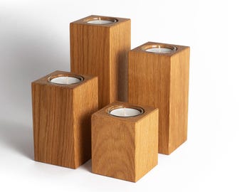 May include: Four wooden candle holders of varying heights. Each holder is a square block with a recessed top to hold a white candle. The wood has a natural grain and a warm, light brown color.