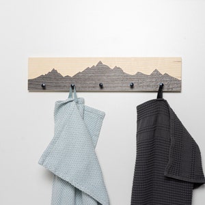 K&ouml;nnte beinhalten: Ein Wand-Kleiderhaken aus Holz mit einem Berg-Silhouetten-Design. Der Haken hat f&uuml;nf Haken und ist in einem hellbraunen und dunkelgrauen Farbschema lackiert. Zwei Handt&uuml;cher h&auml;ngen an den Haken, eines hellblau und eines dunkelgrau.