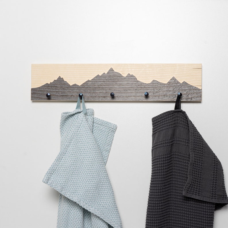 K&ouml;nnte beinhalten: Ein Wand-Kleiderhaken aus Holz mit einem Berg-Silhouetten-Design. Der Haken hat f&uuml;nf Haken und ist in einem hellbraunen und dunkelgrauen Farbschema lackiert. Zwei Handt&uuml;cher h&auml;ngen an den Haken, eines hellblau und eines dunkelgrau.