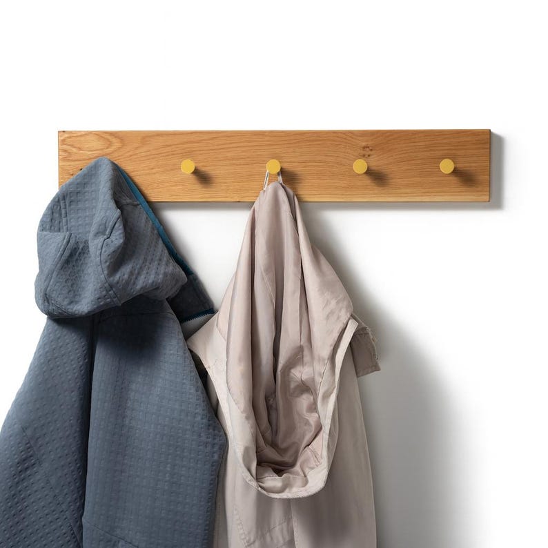 Op de afbeelding: Een houten wandkapstok met vier gele haken. Twee jassen met capuchon, een blauwe en een beige, hangen aan de haken. De kapstok is gemaakt van lichtgekleurd hout en is aan een witte muur bevestigd.