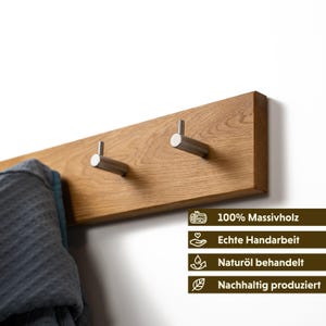 Op de afbeelding: Een houten wandkapstok met twee roestvrijstalen haken. De kapstok is gemaakt van lichtbruin hout en is bevestigd aan een witte muur. Een grijze badjas hangt aan de linkerhaak. De kapstok heeft tekst die zegt "100% Massivholz", "Echte Handarbeit", "Natur&ouml;l behandelt" en "Nachhaltig produziert".