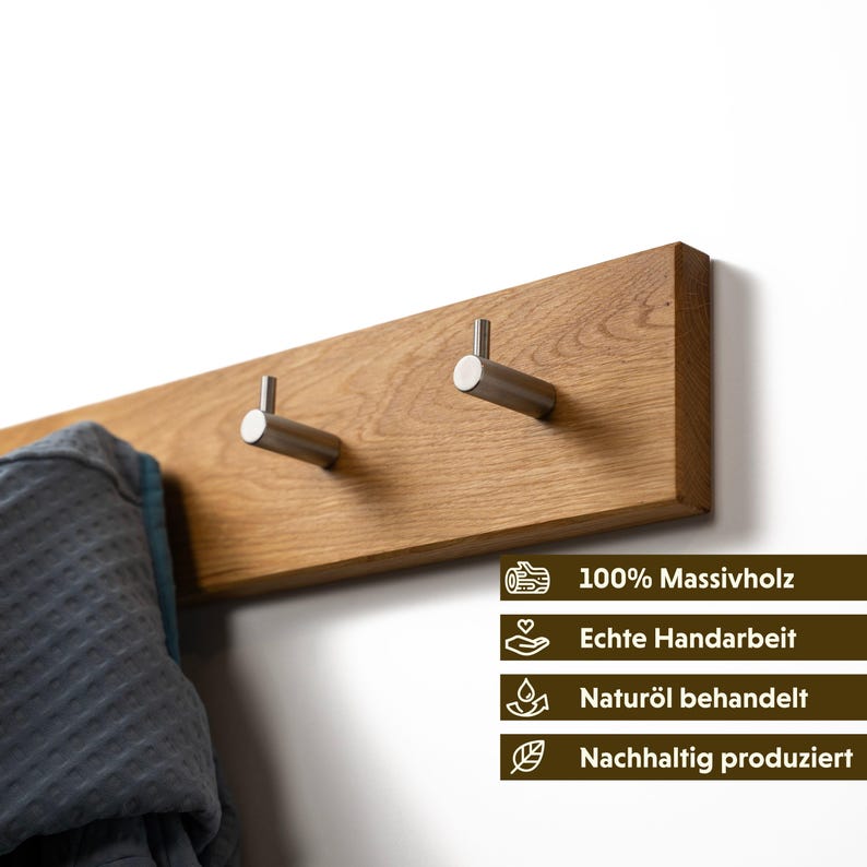 Op de afbeelding: Een houten wandkapstok met twee roestvrijstalen haken. De kapstok is gemaakt van lichtbruin hout en is bevestigd aan een witte muur. Een grijze badjas hangt aan de linkerhaak. De kapstok heeft tekst die zegt "100% Massivholz", "Echte Handarbeit", "Natur&ouml;l behandelt" en "Nachhaltig produziert".