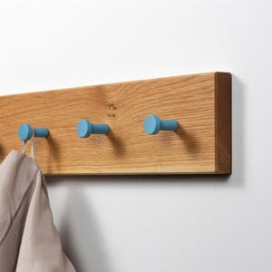Op de afbeelding: Een houten kapstok met drie lichtblauwe haken. De kapstok is gemaakt van lichtbruin hout en is aan een witte muur bevestigd. Een beige kledingstuk hangt aan een van de haken.