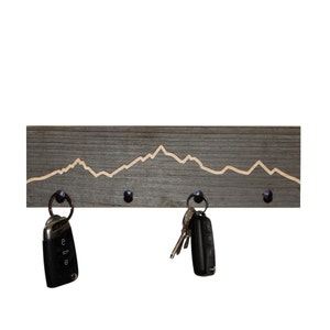 K&ouml;nnte beinhalten: Ein Schl&uuml;sselhalter aus Holz mit einem Bergketten-Design. Der Halter hat vier Haken und ist in Grau lackiert. Zwei Schl&uuml;ssels&auml;tze h&auml;ngen an den Haken.