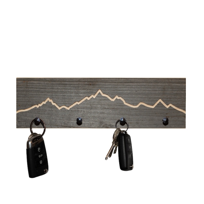 K&ouml;nnte beinhalten: Ein Schl&uuml;sselhalter aus Holz mit einem Bergketten-Design. Der Halter hat vier Haken und ist in Grau lackiert. Zwei Schl&uuml;ssels&auml;tze h&auml;ngen an den Haken.