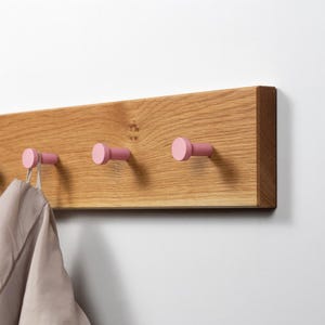 Op de afbeelding: Een houten wandkapstok met drie roze haken. De kapstok is gemaakt van lichtgekleurd hout met een natuurlijke nerf. Een lichtgekleurd kledingstuk hangt aan een van de haken. De haken zijn cilindrisch en geschilderd in een stoffige roze kleur.