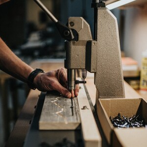 K&ouml;nnte beinhalten: Eine Person verwendet eine Metallpresse, um kleine Metalln&auml;gel an ein Holzbrett zu befestigen. Im Vordergrund ist eine Schachtel mit N&auml;geln zu sehen.