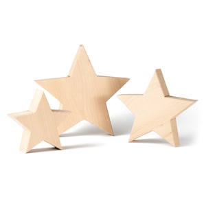 Può includere: Tre stelle di legno di diverse dimensioni. Le stelle sono in legno chiaro non rifinito, che mostra la venatura naturale. Sono disposte su uno sfondo bianco, con la stella più grande al centro.