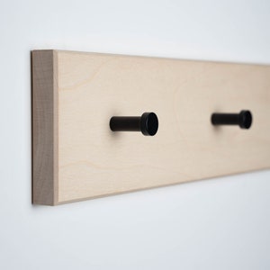 Könnte beinhalten: Ein helles Wandgarderobenbrett aus Holz mit zwei schwarzen zylindrischen Haken. Das Holz hat eine natürliche Maserung, und die Haken sind gleichmäßig verteilt. Das Brett ist rechteckig und an einer weißen Wand montiert.