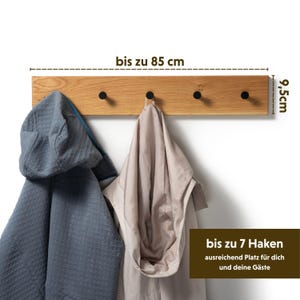 Op de afbeelding: Houten kapstok met vier zwarte haken. De kapstok is 85 cm lang en 9,5 cm hoog. Twee jassen hangen aan de haken. Tekst op de afbeelding luidt "tot 7 haken" en "voldoende ruimte voor jou en je gasten".