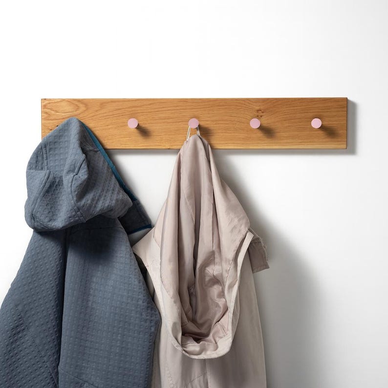 Op de afbeelding: Een houten wandkapstok met vier roze knoppen. Twee jassen, een blauwe en een beige, hangen aan de knoppen. De kapstok is gemaakt van lichtgekleurd hout en is aan een witte muur bevestigd.