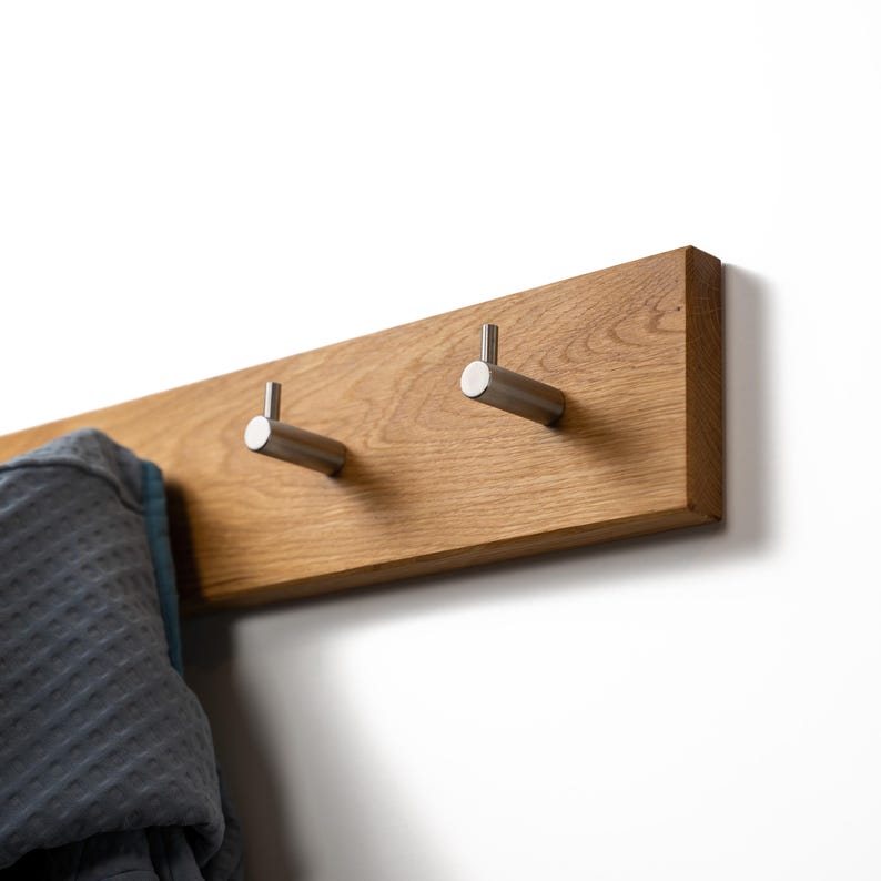Op de afbeelding: Een houten wandkapstok met twee haken van geborsteld nikkel. De kapstok is gemaakt van lichtbruin hout en is aan een witte muur bevestigd. Een grijze badjas hangt aan een van de haken. Modern en minimalistisch ontwerp.