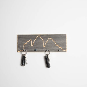K&ouml;nnte beinhalten: Ein grauer Holzschl&uuml;sselhalter mit einem in die Oberfl&auml;che geschnitzten Bergketten-Design. Zwei Schl&uuml;ssel h&auml;ngen an dem Halter.
