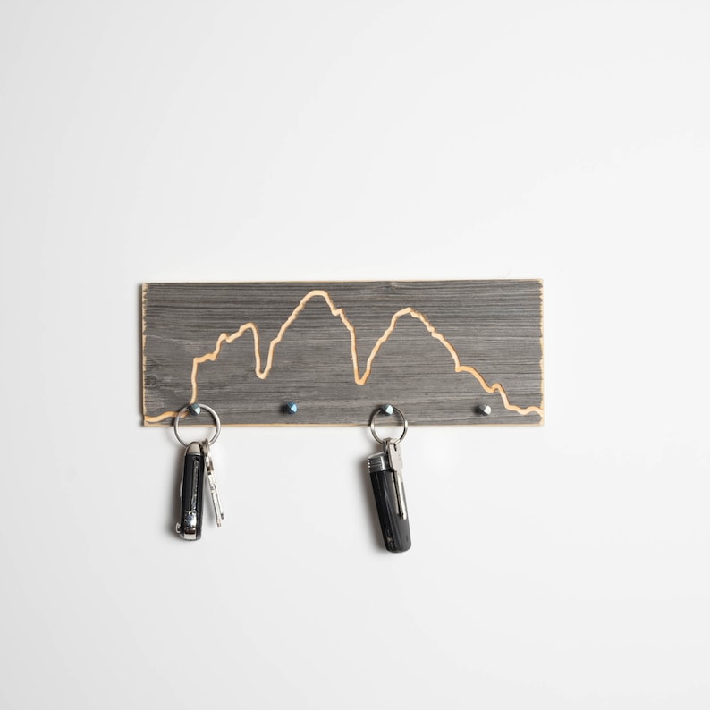 K&ouml;nnte beinhalten: Ein grauer Holzschl&uuml;sselhalter mit einem in die Oberfl&auml;che geschnitzten Bergketten-Design. Zwei Schl&uuml;ssel h&auml;ngen an dem Halter.