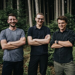 Op de afbeelding: Drie mannen poseren in een bos, met gekruiste armen. De man links draagt een grijs t-shirt, de man in het midden een marineblauwe poloshirt en de man rechts een donker overhemd. Op de achtergrond hoge bomen.