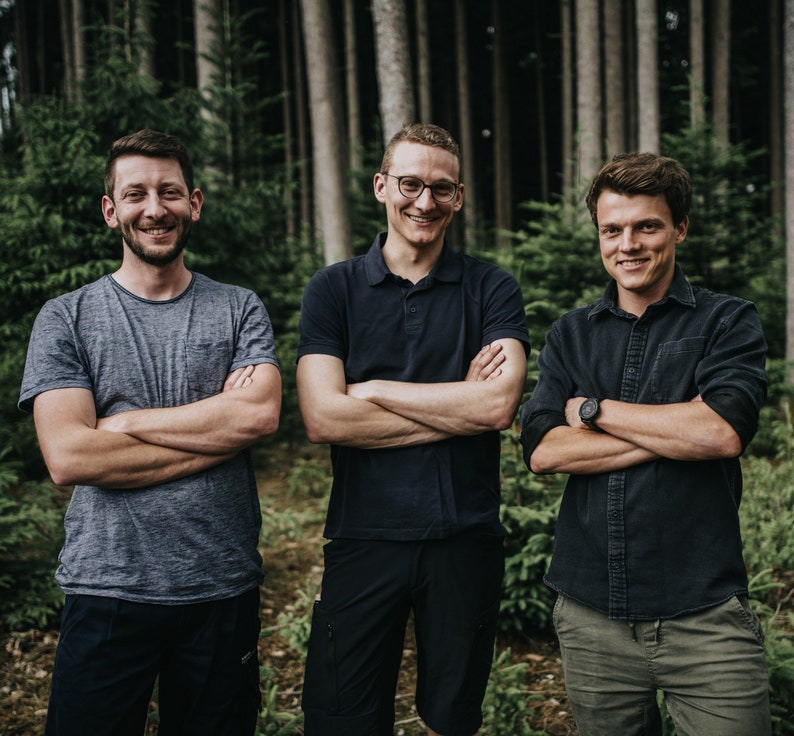 Op de afbeelding: Drie mannen poseren in een bos, met gekruiste armen. De man links draagt een grijs t-shirt, de man in het midden een marineblauwe poloshirt en de man rechts een donker overhemd. Op de achtergrond hoge bomen.
