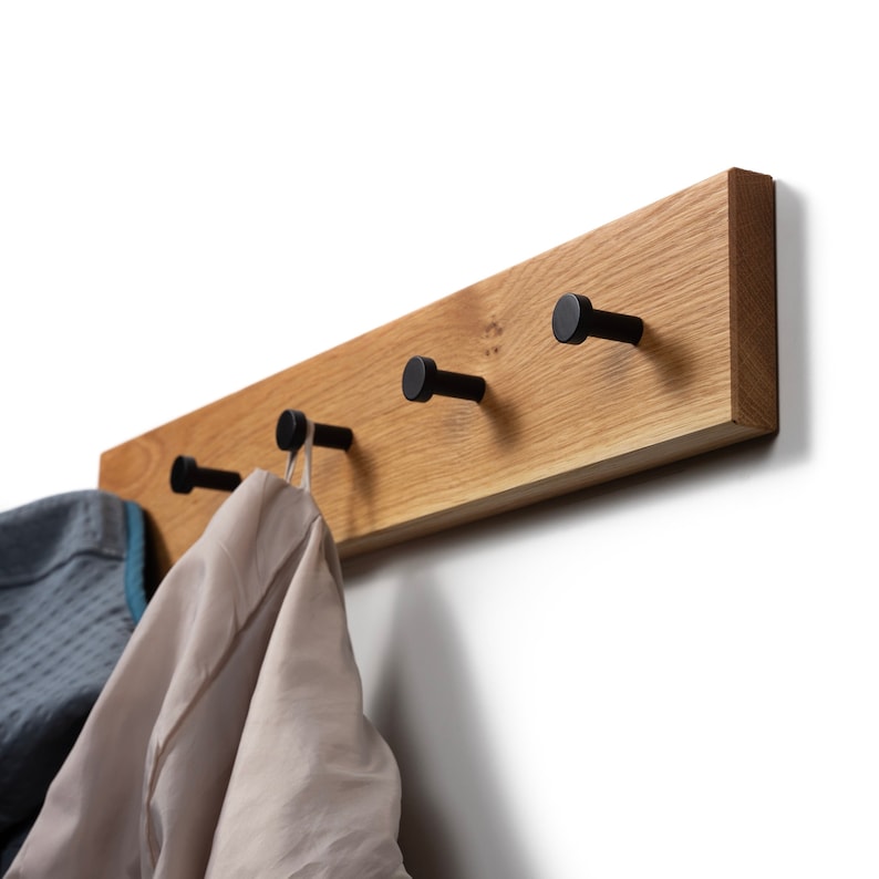Op de afbeelding: Een houten kapstok met vier zwarte metalen haken. De kapstok is gemaakt van lichtgekleurd hout en is aan een witte muur bevestigd. Twee kledingstukken hangen aan de haken. Het ontwerp is minimalistisch en modern.