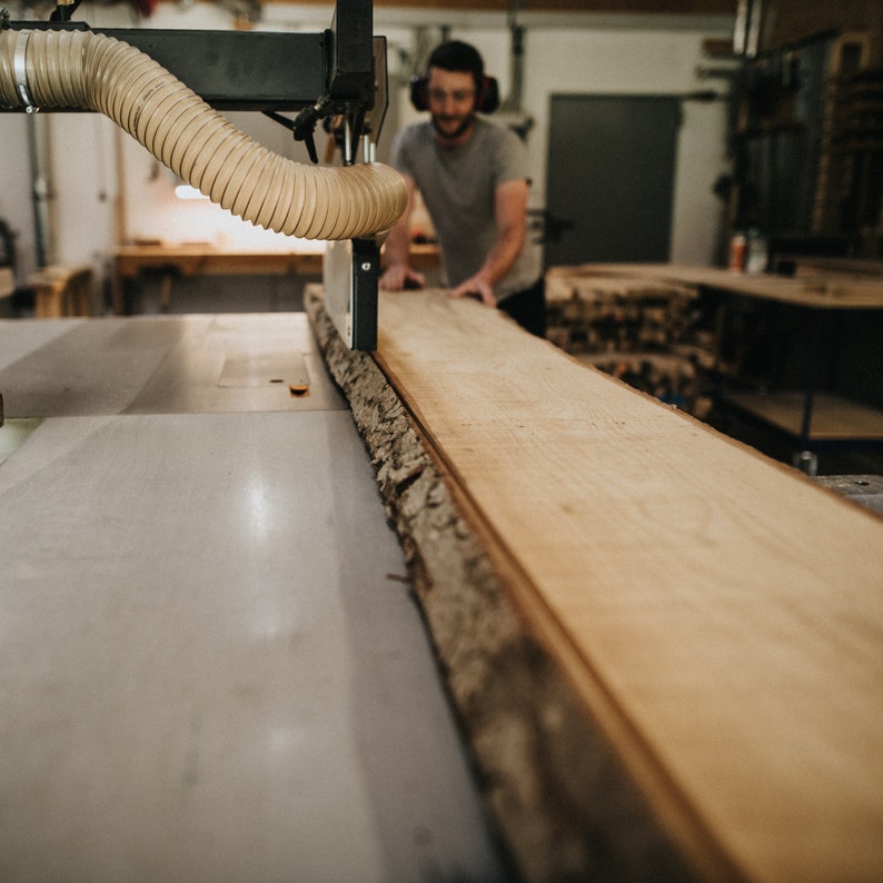 Op de afbeelding: Een timmerman gebruikt een grote zaag om een lange, onafgewerkte houten plank te zagen. Het hout heeft een natuurlijke rand. De werkplaats heeft een neutraal kleurenpalet, met de nadruk op het hout en de machines.