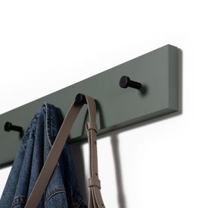 Peut inclure: Un porte-manteau mural en bois vert sauge avec trois crochets en métal noir. Une veste en jean et un sac à main marron clair sont suspendus aux crochets. Le porte-manteau est fixé sur un mur blanc.