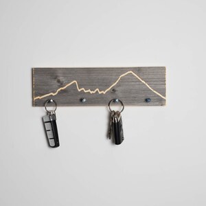 K&ouml;nnte beinhalten: Ein grauer Holz-Schl&uuml;sselhalter mit einem in das Holz geschnitzten Bergketten-Design. Zwei Schl&uuml;ssels&auml;tze h&auml;ngen an dem Halter.