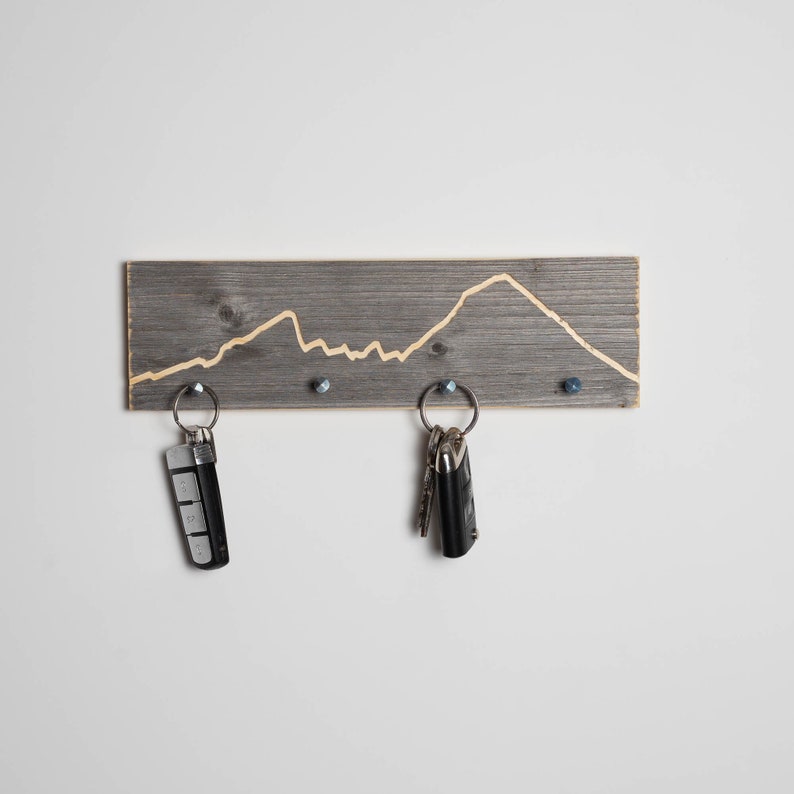 K&ouml;nnte beinhalten: Ein grauer Holz-Schl&uuml;sselhalter mit einem in das Holz geschnitzten Bergketten-Design. Zwei Schl&uuml;ssels&auml;tze h&auml;ngen an dem Halter.