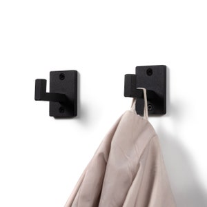 Op de afbeelding: Twee zwarte metalen haken gemonteerd aan een witte muur. Aan één haak hangt een beige jas.