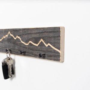 K&ouml;nnte beinhalten: Ein rustikaler Schl&uuml;sselhalter aus Holz mit einem in das Holz geschnitzten Bergketten-Design. Der Halter hat drei schwarze Metallhaken zum Aufh&auml;ngen von Schl&uuml;sseln.