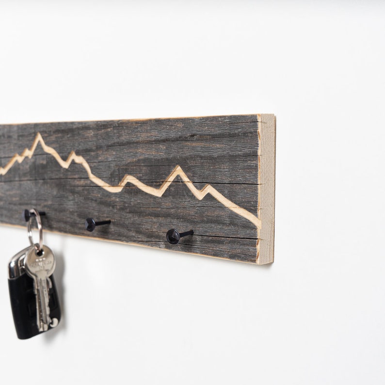 K&ouml;nnte beinhalten: Ein rustikaler Schl&uuml;sselhalter aus Holz mit einem in das Holz geschnitzten Bergketten-Design. Der Halter hat drei schwarze Metallhaken zum Aufh&auml;ngen von Schl&uuml;sseln.