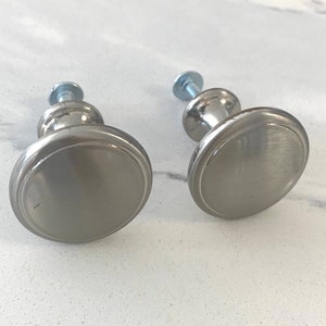 May include: Two brushed nickel cabinet knobs with a flat, oval face and a screw for installation. The knobs are a cool grey colour and have a simple, modern design. They are set against a white marble-patterned surface.