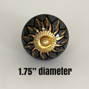 Peut inclure: Bouton de meuble décoratif avec une base noire et un motif floral doré. Le bouton a un diamètre de 4,4 cm. Le centre du bouton est doré et le motif floral est souligné en or.