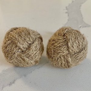 Może przedstawiać: Dwa okrągłe, dekoracyjne węzły z liny. Węzły wykonane są z naturalnej juty, ciasno splecionych w kształt kuli. Węzły leżą na białej i szarej powierzchni.