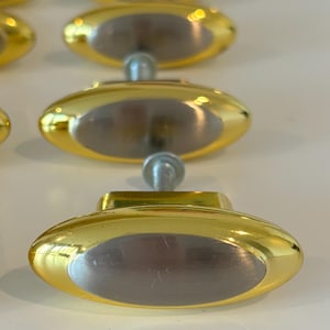 Op de afbeelding: Een verzameling goudkleurige ovale kastknoppen met een zilverkleurig midden. De knoppen zijn in rijen gerangschikt en benadrukken hun gepolijste afwerking en eenvoudige ontwerp. De hardware is waarschijnlijk voor meubels of kasten.