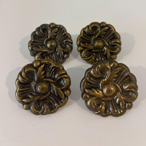 May include: Four antique brass-colored cabinet knobs with a floral design. Each knob features a detailed, raised pattern of leaves and petals, with a central rounded element. The knobs are arranged on a white surface, showcasing their intricate, vintage aesthetic.