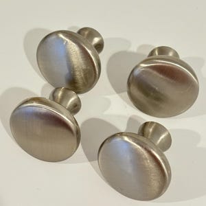 May include: Four brushed nickel cabinet knobs with a rounded, oval design. Each knob has a short, cylindrical stem. The knobs are arranged on a white surface, providing a clean, modern aesthetic. These knobs are suitable for drawers and cabinets.