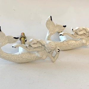 May include: Two off-white mermaid cabinet knobs. The mermaids are sculpted with detailed features, including flowing hair and a tail. Black accents highlight the eyes, fingertips, and tail tips. Each knob includes a brass-colored bolt for installation.