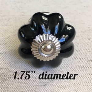 May include: A black ceramic cabinet knob with a silver centre and a flower-like design. The knob has a diameter of 4.4 cm.