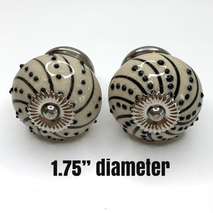 May include: Two decorative cabinet knobs with a cream-colored base, adorned with black spiral and dot patterns. Each knob features a silver-colored metal base and a decorative silver-colored flower-like center. The text "1.75" diameter" is visible.