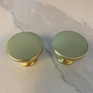 Puede incluir: Dos tiradores dorados para armarios con acabado cepillado. Cada tirador tiene una parte superior redonda y una base cilíndrica corta. Los tiradores están sobre una superficie con vetas blancas y grises.